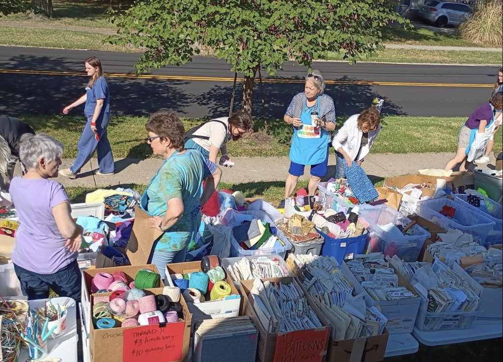 Volunteers and shoppers mingle around tables of craft supplies at the Garth location during the annual supplies yard sale. Volunteers wear bright blue aprons.